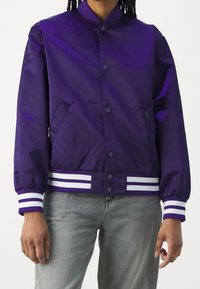 Paarse bomberjack met glanzende afwerking, gebreide witte en paarse gestreepte manchetten en zoom, en twee zijzakken. Knopenluiting aan de voorkant.