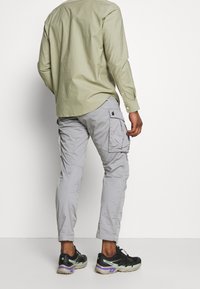 Hombre vestido con una camiseta de manga larga verde claro y pantalones cargo grises claros, con zapatillas Salomon negras y beige, de pie frente a una pared blanca lisa.