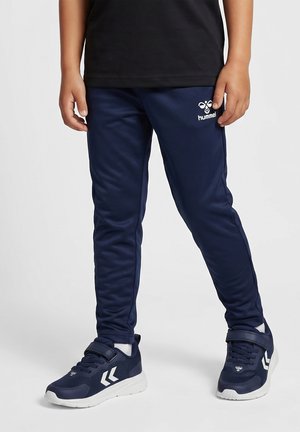 Kind draagt marineblauwe Hummel trainingsbroek en bijpassende sneakers met witte zolen en logo's, staande tegen een effen achtergrond.