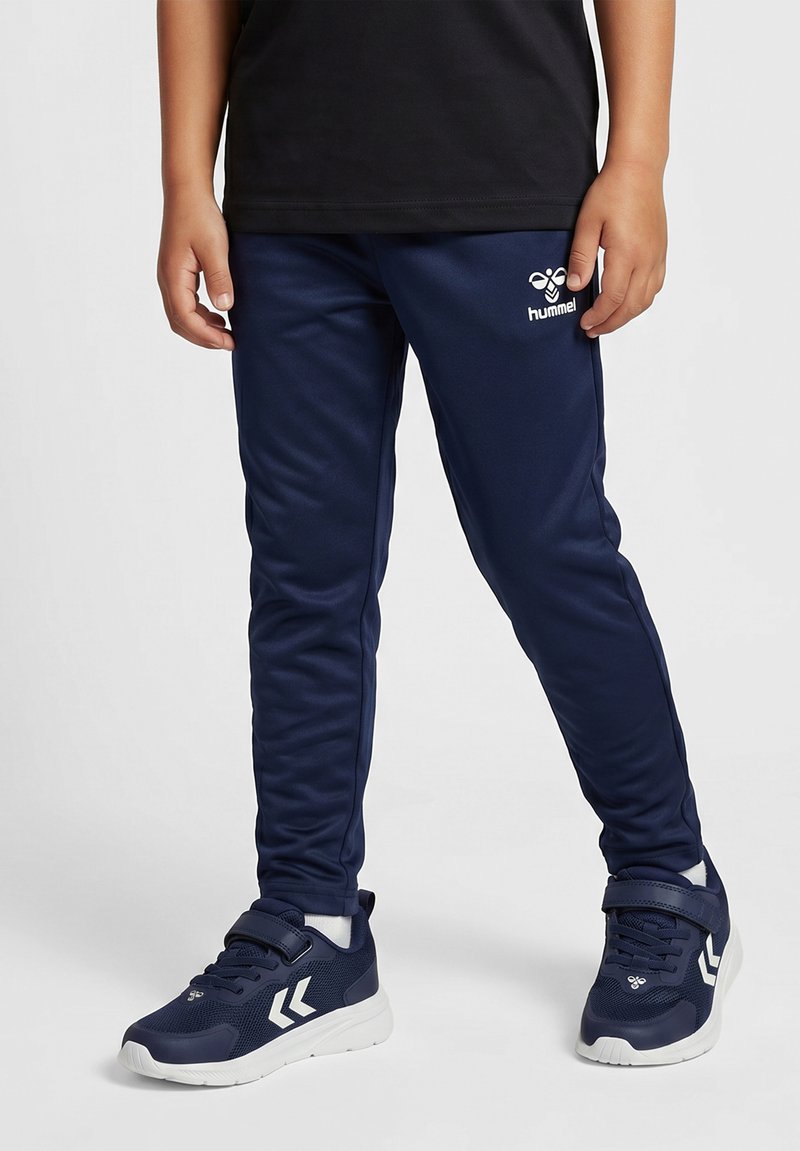 Dziecko ubrane w granatowe spodnie dresowe marki Hummel oraz pasujące buty sportowe z białymi podeszwami i logo, stojące na jednolitym tle.
