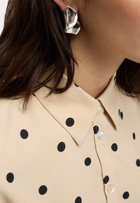 Beigefärgad skjorta med svarta prickar, med klassisk krage och slät yta. Silverfärgad statementöronprydnad synlig bredvid kragen.