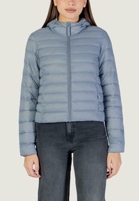 Giacca imbottita trapuntata azzurro chiaro con cappuccio, cerniera frontale e polsini elastici, abbinata a jeans scuri. Tessuto morbido in tutta la giacca.