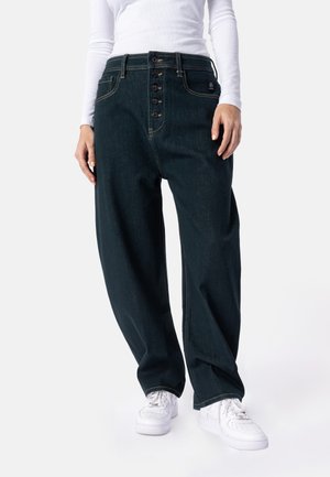 Person trägt weite dunkelblaue Jeans mit fünf sichtbaren Knöpfen, ein weißes Langarmshirt und weiße Sneakers und steht aufrecht.