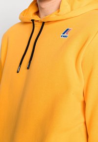 Żółta bluza z kapturem, z czarnymi sznurkami, logo K-WAY w niebieskim i czerwonym na klatce piersiowej, wykonana z miękkiego materiału, z kieszenią kangurzą.
