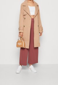 Beżowy płaszcz trencz z dwurzędowym zapięciem, szerokie brązowe culottes, biały krótki top, białe trampki z różowymi akcentami oraz mała beżowa torebka.