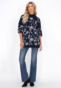 Marineblauwe pullover trui met witte bloemenborduring, losse fit, lange mouwen en een zachte textuur. Gecombineerd met flared denim jeans en zwarte laarzen.
