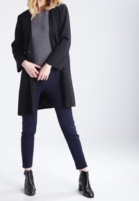 Černý dlouhý kabát s texturovaným vzorem, šedý svetr, tmavě modré úzké kalhoty a černé kotníkové boty s nízkým podpatkem.