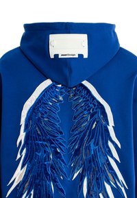 Sudadera azul con grandes alas bordadas en azul oscuro y acentos en blanco. Incluye una etiqueta de tela en la parte posterior de la capucha.