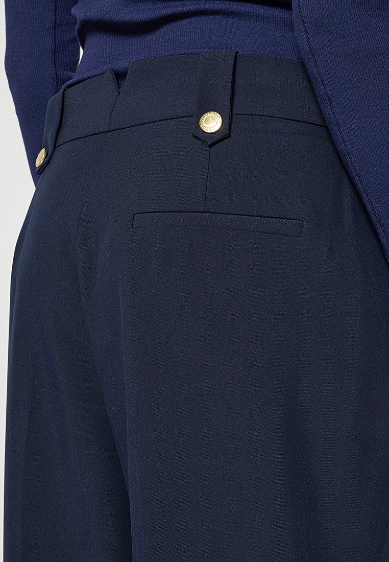 Pantalon bleu marine ajusté avec une poche passepoilée à l'arrière et des boutons décoratifs dorés sur les passants de ceinture, porté avec un haut assorti bleu marine.