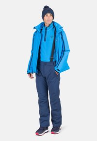 Giacca impermeabile blu con polsini regolabili, strato intermedio in pile blu, pantaloni isolati blu navy con bretelle e calzature scure con dettagli rossi.