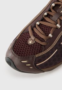 Shoe de desporto castanho com parte superior em malha, sola de borracha e realces em castanho claro contrastantes. Texturas distintas com fecho de atacadores.