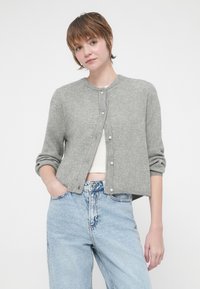 Cardigã cinza com nervuras, mangas longas, decote redondo e fecho frontal com botões. Usado sobre uma blusa branca e combinado com jeans azuis claros.