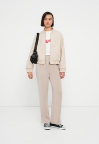 Μπεζ μπουφάν bomber πάνω από ένα λευκό μπλουζάκι με κόκκινο γραφικό, σετ με ανοιχτό μπεζ παντελόνι και μαύρα αθλητικά παπούτσια. Συμπεριλαμβάνεται μαύρη τσάντα ώμου.