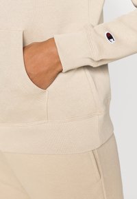 Μπεζ φούτερ με μπροστινή τσέπη και ριμπ μανσέτες. Διαθέτει μικρό κεντημένο λογότυπο στο μανίκι. Κατασκευασμένο από μαλακό ύφασμα.