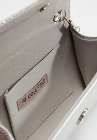 Interior de un bolso de mano plateado con un forro de tela suave, un bolsillo para tarjetas etiquetado "ANNA FIELD" y un cierre metálico circular.