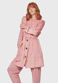 Lunga veste rosa in morbido tessuto, con vita avvolgente, scollatura profonda a V e grandi bottoni. Abbinata a pantaloni rosa coordinati.