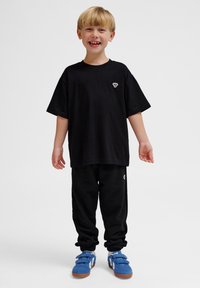 Schwarzes Baumwoll-T-Shirt mit kurzen Ärmeln und kleinem Logo, kombiniert mit schwarzen Chino-Hosen und blauen Turnschuhen mit Klettverschluss.