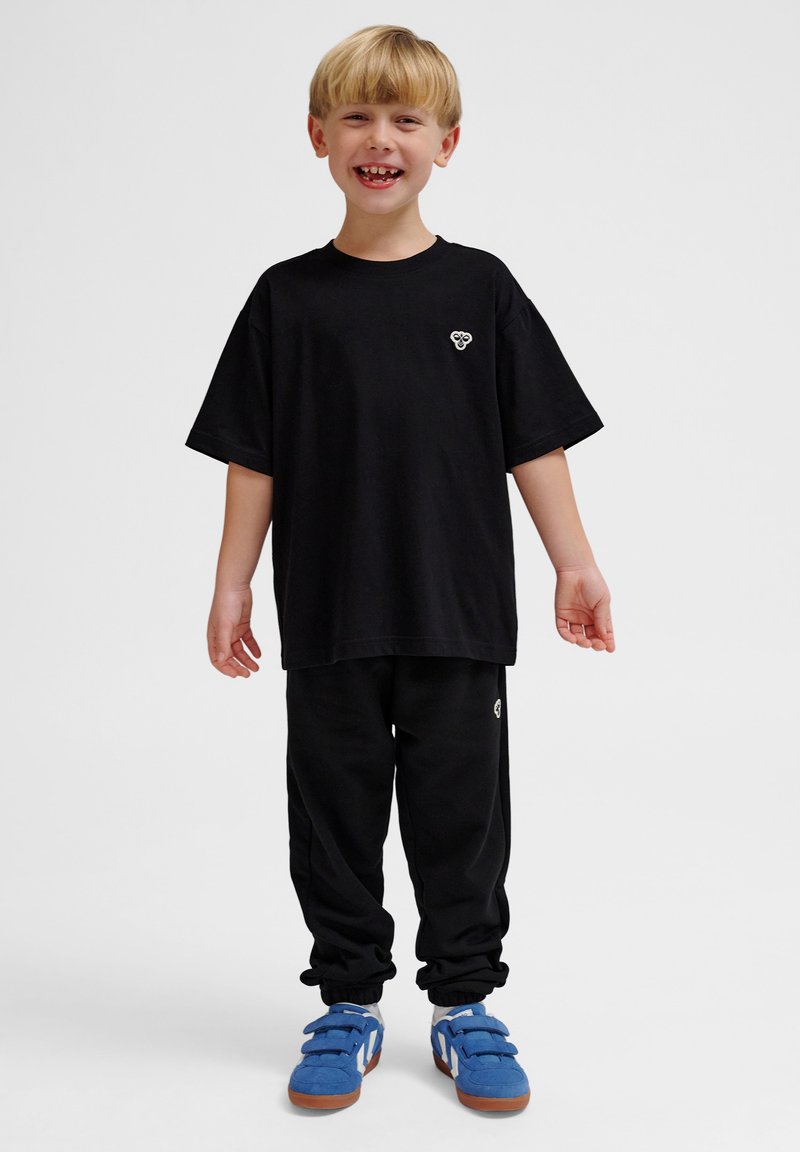 Schwarzes Baumwoll-T-Shirt mit kurzen Ärmeln und kleinem Logo, kombiniert mit schwarzen Chino-Hosen und blauen Turnschuhen mit Klettverschluss.