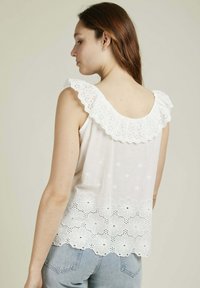 Haut sans manches blanc avec un col en eyelet volanté, broderies florales sur l'ourlet, et une coupe ample, porté avec un jean bleu clair.