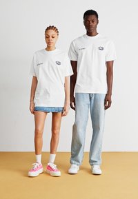 Duas pessoas vestem t-shirts de algodão brancas com designs gráficos. A modelo feminina combina a sua com uma saia de ganga e sapatilhas cor-de-rosa.