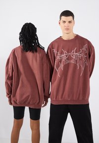 Dos personas visten sudaderas burdeos de gran tamaño. En la parte delantera presentan un diseño gráfico en blanco. La tela parece suave y tiene un corte holgado.
