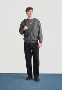 Γκρι φούτερ με φερμουάρ και μπροστινές τσέπες, φορεμένο πάνω από γκρι μπλουζάκι, συνδυασμένο με μαύρα τζιν παντελόνια και σκούρα παπούτσια. Μικρή πράσινη τσάντα φορεμένη στον ώμο.