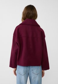 Burgundowa wełniana kurtka w oversize z szerokimi klapami, opadającymi ramionami i prostym dołem, noszona na jasnoniebieskich dżinsowych spodniach.