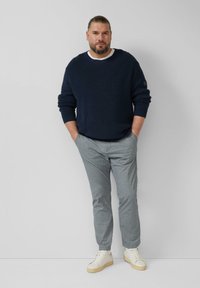 Marineblauwe geribde trui met witte kraag, lichtgrijze broek met patroon en witte sneakers. Man staat nonchalant met handen in de zakken.