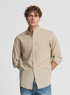 Młody mężczyzna z kręconymi włosami ubrany w beżową koszulę na guziki z podwiniętymi rękawami oraz niebieskie jeansy, stojący na jednolitym białym tle.