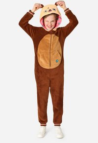 Rjav flis onesie z bež trebuhom, kapuco v obliki živali z ušesi in obrazom, spredaj z zadrgo in stranskimi žepi. Na nogah so bele športne copate.