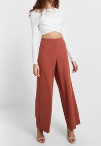Femme portant un pantalon taille haute, large, de couleur rouille, un crop top torsadé à manches longues blanc et des talons ouverts beige.