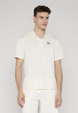 Ung mann iført en tettsittende hvit polotrøye og hvite bukser, stående mot en ensfarget lys grå bakgrunn, vendt litt mot høyre.