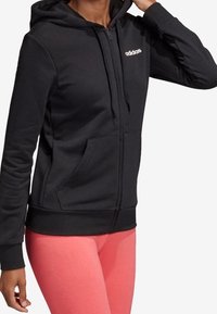 Sweat à capuche zippé noir en tissu doux, avec une poche avant et une capuche ajustable. Logo blanc sur la poitrine. Associé à des leggings roses.