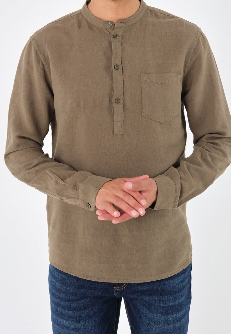 Camicia marrone a maniche lunghe con colletto alla cinese, apertura a bottoni e taschino sul petto. Il materiale sembra morbido con una finitura testurizzata.