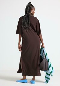 Brun maxikjole med korte ærmer, sideslids og en afslappet pasform. Sammen med blå flade sko og holder en stribet tæppe og en håndtaske.