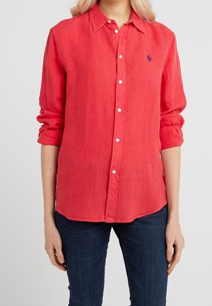 Femme portant une chemise rouge vif à manches longues avec les manches retroussées et un jean bleu foncé sur un fond uni.