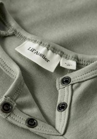 Grön bomullsplagg med tryckknappar och rundad halsringning. Har mjuk textur och stuckna kanter. Etiketten säger "Lil' Atelier."