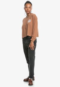 Bruine cropped lange mouwen shirt met grafisch ontwerp, gecombineerd met zwarte high-waisted jeans en bruine schoenen, gefotografeerd tegen een witte achtergrond.