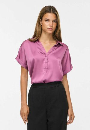 Femme portant un chemisier violet en satin à manches courtes avec boutons et un pantalon noir taille haute sur un fond blanc uni.