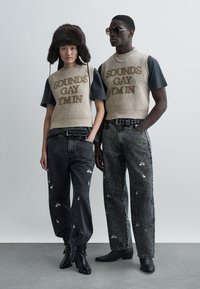 Dwie modelki stoją obok siebie. Jedna ma na sobie beżowy dzianinowy top z brązowym napisem oraz czarne dżinsy z białymi wzorami; druga ubrana jest w czarną koszulę i dżinsy.