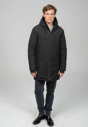 Schwarzer gepolsterter Wintermantel mit einer Kordelzugkapuze, Reißverschluss, zwei seitlichen Taschen und mittellanger Länge; getragen mit dunklen Jeans und Stiefeln.