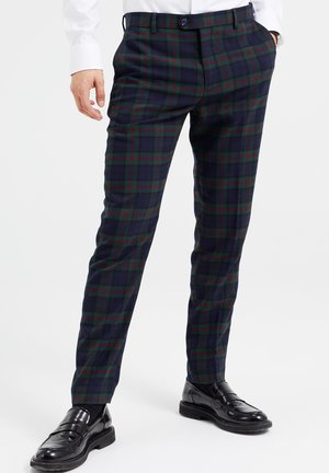 Man draagt marineblauwe, groene en rode geruite slim-fit broek met zwarte leren loafers en witte blouse, handen in de zakken.