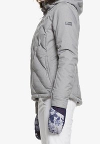 Graue isolierte Jacke mit Kapuze, die ein Rautenmuster aufweist. Kombiniert mit marineblauen Handschuhen, die ein florales Design zeigen.