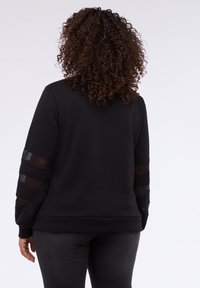 Mujer con cabello rizado que lleva una camiseta de manga larga negra con franjas de malla transparente en las mangas y pantalones oscuros ajustados, de pie contra un fondo blanco.