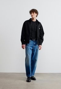 Νεαρός άνδρας που στέκεται με μαύρο μπουφάν, μαύρο πουκάμισο, μπλε τζιν και μαύρα παπούτσια μπροστά από απλό ανοιχτό γκρι φόντο.