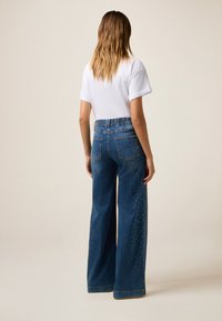 Denim wijdelaars met een hoge tailleband, voorzien van decoratieve studs op de benen en een vervaagde blauwe wash. Achterzakken zichtbaar.