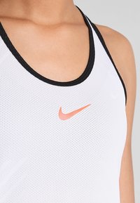 Top atletico bianco con rifiniture nere, caratterizzato da una struttura in rete e un logo Nike arancione. Il design sottolinea la traspirabilità e il comfort.