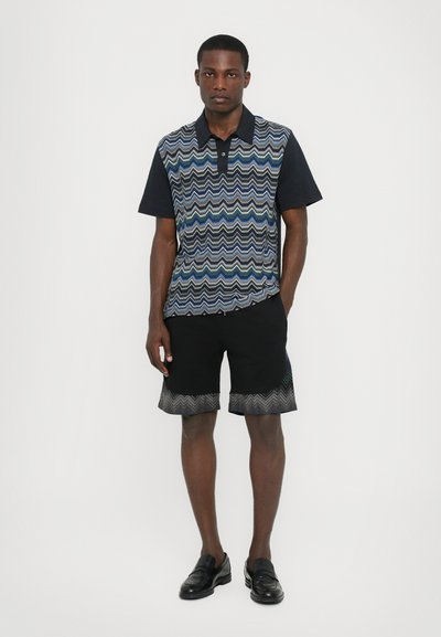 Polo cu mânecă scurtă de culoare bleumarin, cu un model zigzag în nuanțe de albastru și verde, asortat cu pantaloni scurți negri ce au un detaliu zigzag în aceeași nuanță.