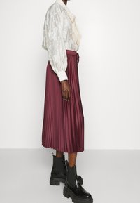 Blusa bianca texturizzata con un fiocco al collo, abbinata a una gonna plissettata bordeaux. Stivaletti neri alla caviglia completano l'outfit.