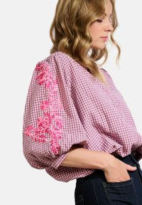 Blusa a scacchi rosa con maniche a puff, decorata con intricati ricami floreali rosa sulla manica sinistra e dettagli con bottoni sul davanti.